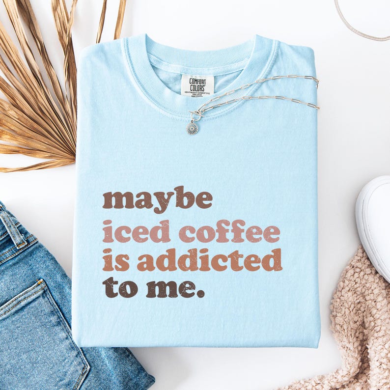 Peut inclure: T-shirt bleu clair pli&eacute; avec l'inscription "maybe iced coffee is addicted to me" en marron et rose. Le t-shirt est orn&eacute; d'un collier argent&eacute; avec un petit pendentif. Le t-shirt est de la marque Comfort Colors.
