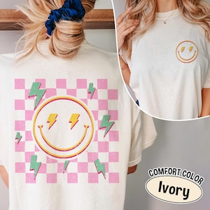 May include: Ivory-colored t-shirt featuring a smiley face with lightning bolts and a pink and white checkered background. The shirt also has a smaller smiley face with lightning bolts on the front. The text "COMFORT COLOR Ivory" is displayed.