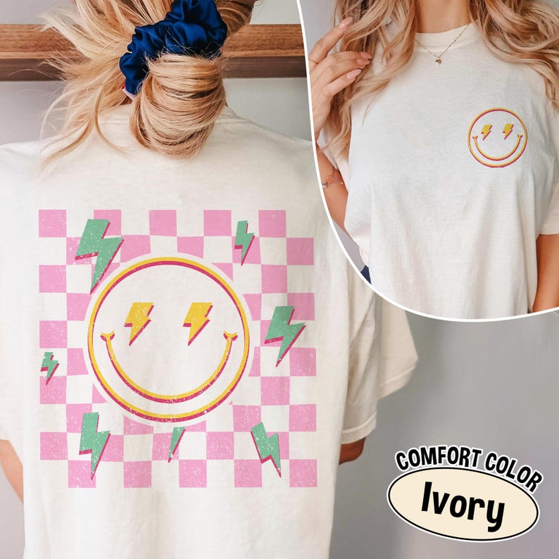 May include: Ivory-colored t-shirt featuring a smiley face with lightning bolts and a pink and white checkered background. The shirt also has a smaller smiley face with lightning bolts on the front. The text "COMFORT COLOR Ivory" is displayed.