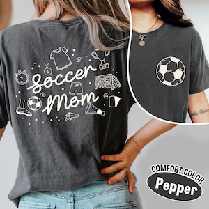 K&ouml;nnte beinhalten: Dunkelgraues Comfort Color T-Shirt mit einem wei&szlig;en Fu&szlig;ball-Grafik auf der Vorderseite und einem "Soccer Mom"-Design auf der R&uuml;ckseite. Das R&uuml;ckendesign enth&auml;lt fu&szlig;ballbezogene Illustrationen. Das Shirt besteht aus einem weichen, bequemen Material.