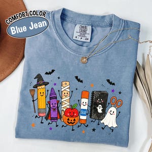 May include: A blue jean colored t-shirt featuring a graphic print of anthropomorphic school supplies dressed for Halloween, including a pencil witch, a crayon vampire, a mummy ruler, a pumpkin, a glue stick, a ghost notebook, and ghost scissors.