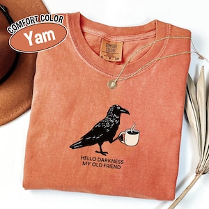 May include: A Comfort Colors Yam-colored t-shirt featuring a black raven holding a coffee cup. The shirt has the text "HELLO DARKNESS MY OLD FRIEND" printed below the raven. A gold necklace is draped across the shirt.
