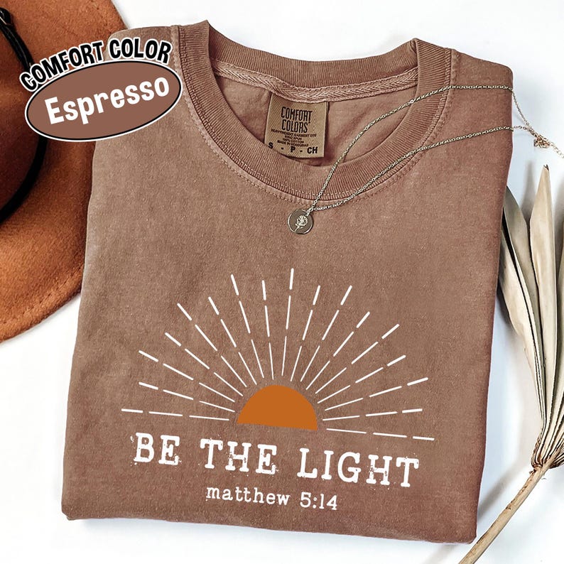 May include: Espresso-colored t-shirt with a white sunburst design and the words "BE THE LIGHT" and "matthew 5:14". The shirt has a crew neck and a small tag. A gold necklace with a pendant is draped across the shirt.