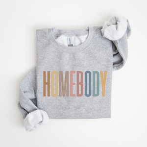 May include: A light gray sweatshirt with the word "HOMEBODY" printed across the front in a retro color palette of brown, beige, pink, blue, and yellow. The sleeves are folded, revealing a white interior.