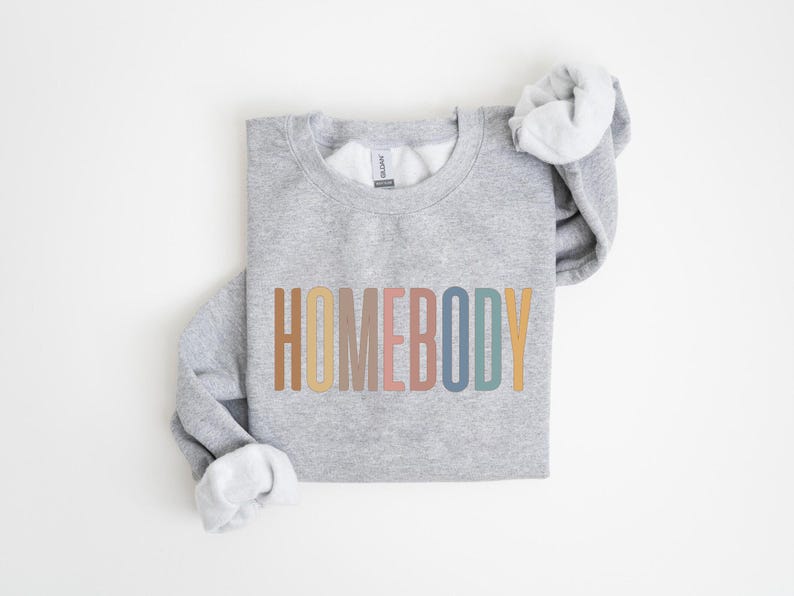 May include: A light gray sweatshirt with the word "HOMEBODY" printed across the front in a retro color palette of brown, beige, pink, blue, and yellow. The sleeves are folded, revealing a white interior.