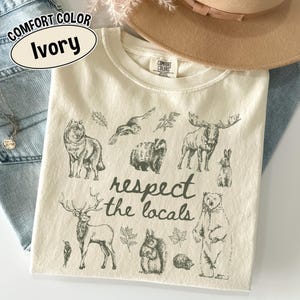 May include: Ivory Comfort Colors t-shirt with woodland animal illustrations, such as a wolf, moose, and bear. The phrase "respect the locals" is printed in black script below the animal drawings.