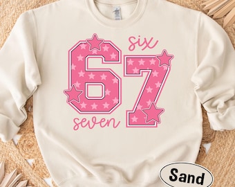 May include: A sand-colored sweatshirt with the number "67" in pink, filled with white stars. The word "six" is above the number, and "seven" is below. Pink star accents are placed around the numbers. A sand-colored oval label at the bottom says "Sand".