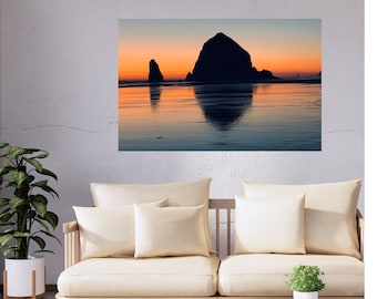 May include: A photograph of a large rock formation silhouetted against a sunset sky, reflected in the calm water of the ocean. The rock formation is in the center of the image, with a smaller rock formation to the left. The sky is a mix of orange, pink, and purple.