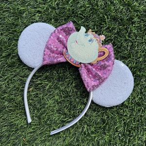May include: A silver headband with white sequined mouse ears. A magenta sequined bow is centered on the headband, adorned with a glittery, light green teapot character. The headband is set against a green grass background.