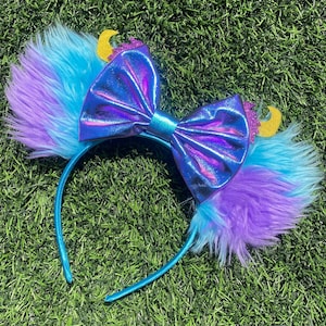May include: A blue headband with faux fur ears in shades of blue and purple. A large, iridescent bow sits between the ears. Gold glitter crescent moon accents are attached to the ears. The headband is on a green surface.