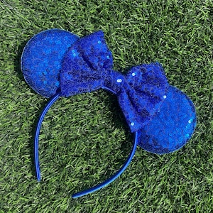 May include: Blue sequined Minnie Mouse ears headband with a large bow. The headband is made of a blue fabric and has a blue bow with a sequined design.