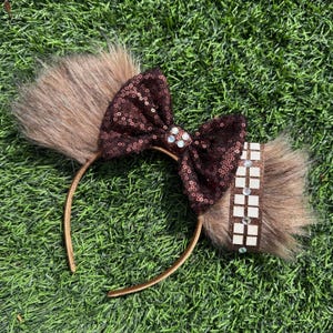 May include: A brown headband with faux fur accents and a sequined bow. The bow is embellished with small, sparkling gems. A brown band with white squares is on the side. The headband is on a green surface.
