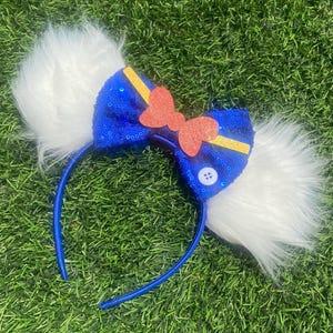 May include: Blue and yellow sequined headband with a red bow and white fur ears. The headband has a white button with a black dot in the center.