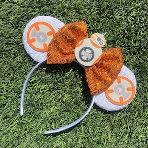 May include: White headband with orange and white ears featuring a white and orange character with a black circle in the center. The headband has a large orange bow with sequins.