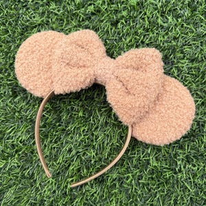 May include: A tan-colored headband with a large, fluffy bow and two round ear-shaped accents. The headband is made of a soft, textured material and has a gold-colored band. The accessory is set against a green background.