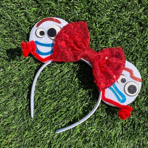 May include: White headband with red sequin bow and two white ears with a cartoon face on each. The ears have blue and red details and glitter accents.