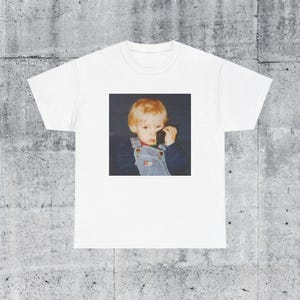 May include: White t-shirt with a photo of a young boy with blonde hair wearing a blue and white overall and holding a black phone to his ear.