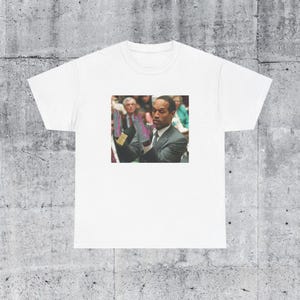 May include: A white t-shirt with a black and white image of a man in a suit with his hands raised. The man is wearing a purple tie and has a serious expression on his face.