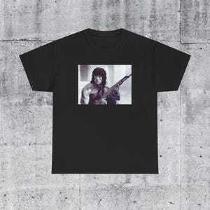 May include: Black t-shirt featuring a graphic of a man holding a rifle. The image is a black and white portrait. The t-shirt is made of a soft, comfortable material. The shirt is a crew neck style.