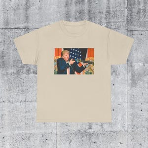 May include: Beige t-shirt featuring a graphic of a person holding a firearm. The graphic includes a person in a suit and tie, with a U.S. flag in the background. The t-shirt is made of a soft, comfortable material.