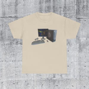 May include: A beige t-shirt with a computer graphic. The graphic shows a monitor displaying a game scene, a computer tower, keyboard, mouse, and a game controller. The design is centrally placed on the shirt.