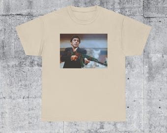 May include: A beige t-shirt with a print of a man holding a gun. The man is wearing a suit and tie. The background is a blurry image of a city street.