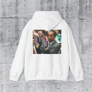 May include: White hoodie featuring a graphic of a man in a suit, with his hands raised, in front of a courtroom background. The image on the hoodie is a photograph of a famous person. The hoodie is made of a soft material.