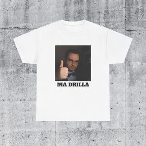 May include: White t-shirt with a black and white photo of a person giving a thumbs up. The text "MA DRILLA" is printed in black below the photo.