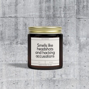 May include: A brown glass candle jar with a gold lid. The label reads "Smells like headshots and hacking accusations" with the text "CSZTHREADS", "4 oz", "20 hours", and "HAND POURED IN USA".