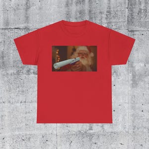 May include: Red t-shirt featuring a rectangular image of Santa Claus holding a toy gun. Santa is wearing a red hat and glasses. The background is blurred, suggesting an indoor setting. The shirt is made of a soft material.