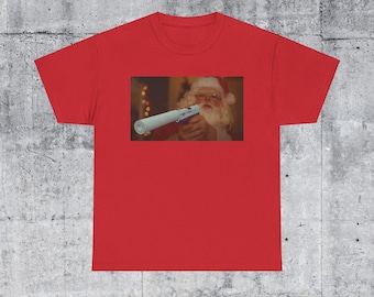 May include: Red t-shirt featuring a rectangular image of Santa Claus holding a toy gun. Santa is wearing a red hat and glasses. The background is blurred, suggesting an indoor setting. The shirt is made of a soft material.