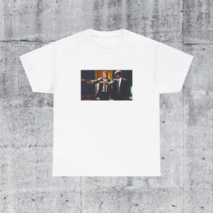 May include: White t-shirt featuring a movie scene with two men in suits, one pointing a gun. The image is a colorful print on the front of the shirt. The t-shirt is made of a soft, comfortable material.