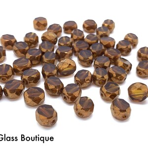 May include: A collection of small, faceted, bronze-colored glass beads scattered across a white surface. Each bead features a metallic finish and a central hole. The text "Bead & Glass Boutique" is printed in black at the bottom left corner.
