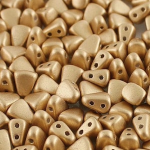 May include: Close-up of a pile of golden-colored, two-hole beads. The beads have a matte finish and a rounded, triangular shape. These beads are ideal for jewelry making and crafting projects.