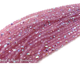 May include: A collection of light pink, faceted glass beads arranged in multiple strands. The beads feature an iridescent finish that reflects light with blue and silver highlights. The text "Bead & Glass Boutique" is printed in white at the bottom.