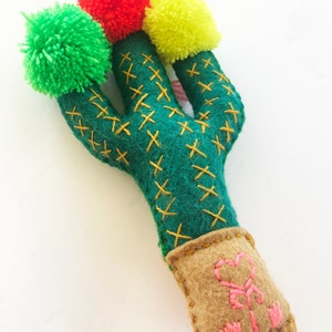 May include: A green felt cactus with yellow and red pom-poms on top. The cactus is in a brown felt pot with a pink heart embroidered on the front.