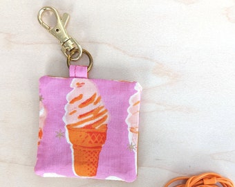 May include: A pink fabric earbud case with a gold clasp, featuring an ice cream cone pattern. Next to it are white earbuds and an orange audio cable.