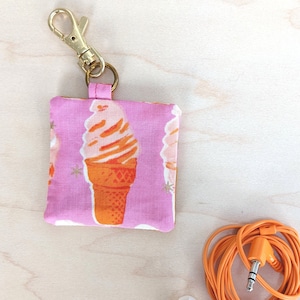 May include: A pink fabric earbud case with a gold clasp, featuring an ice cream cone pattern. Next to it are white earbuds and an orange audio cable.