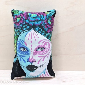 Puede incluir: Una almohada colorida con fondo negro y una cara blanca con maquillaje rosa y azul. La cara tiene una corona floral con flores rosas, azules y verdes.