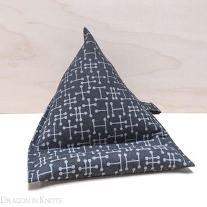 May include: A gray and white patterned triangular pillow with a small loop on the side. The pillow has a geometric design and is made of fabric. The pillow is designed to prop up a tablet or book.