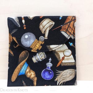 May include: Black fabric pouch with a flap closure, featuring a repeating pattern of wizarding tools and accessories. The design includes potion bottles, a crystal ball, books, and a witch's hat. The pouch is square and has a button closure.