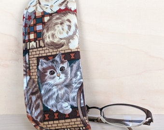 May include: A fabric eyeglasses case with a cat pattern. The case features a repeating design of fluffy gray and brown cats on a tan background with a brick-like pattern. The case is next to a pair of eyeglasses.
