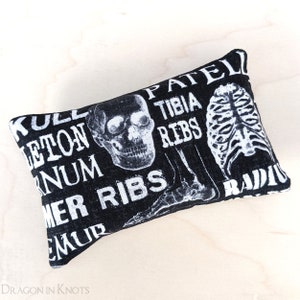 May include: A black and white rectangular pillow featuring a skull and skeletal diagrams. The pillow has text that includes the words: skull, skeleton, sternum, femur, patella, tibia, ribs, and radius. The pillow is a decorative item.