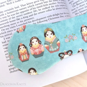 May include: A turquoise fabric bookmark decorated with a pattern of Matryoshka dolls in shades of pink, green, and yellow. The bookmark is positioned on an open book. The text "DRAGON IN KNOTS" is visible at the bottom.
