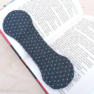 May include: A fabric bookmark with a dark gray background and a repeating pattern of teal hexagons. The bookmark is placed on an open book with printed text.