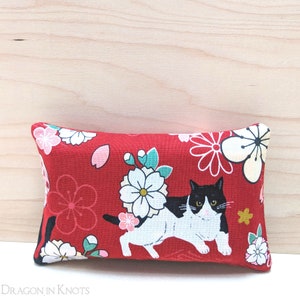 May include: Red fabric cushion with a black and white cat design and white flowers. The cat is facing forward and has a black nose and whiskers.