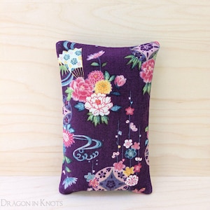 May include: A rectangular, purple pincushion with a floral pattern. The design features pink, white, and yellow flowers, along with blue and white accents. The pincushion is made of fabric and is likely used for sewing or crafting.