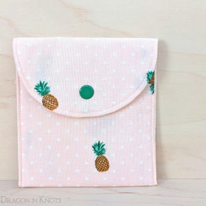 Puede incluir: Una bolsa de tela rosa con cierre de solapa y un broche verde. La bolsa está decorada con lunares blancos y piñas bordadas. La bolsa mide aproximadamente 13 cm de ancho y 13 cm de alto.