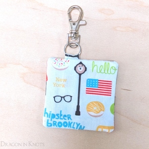 May include: A small white fabric keychain with a New York City theme. It features a donut, an American flag, a street lamp, glasses, and the words "hello", "New York", "hipster", and "Brooklyn".