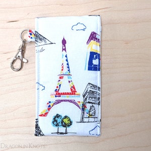 May include: White fabric eyeglasses case with a colorful Eiffel Tower and Parisian landmarks print. Includes a silver-colored clasp and a small loop for attaching to a bag or keychain. The case is rectangular and has a stitched edge.